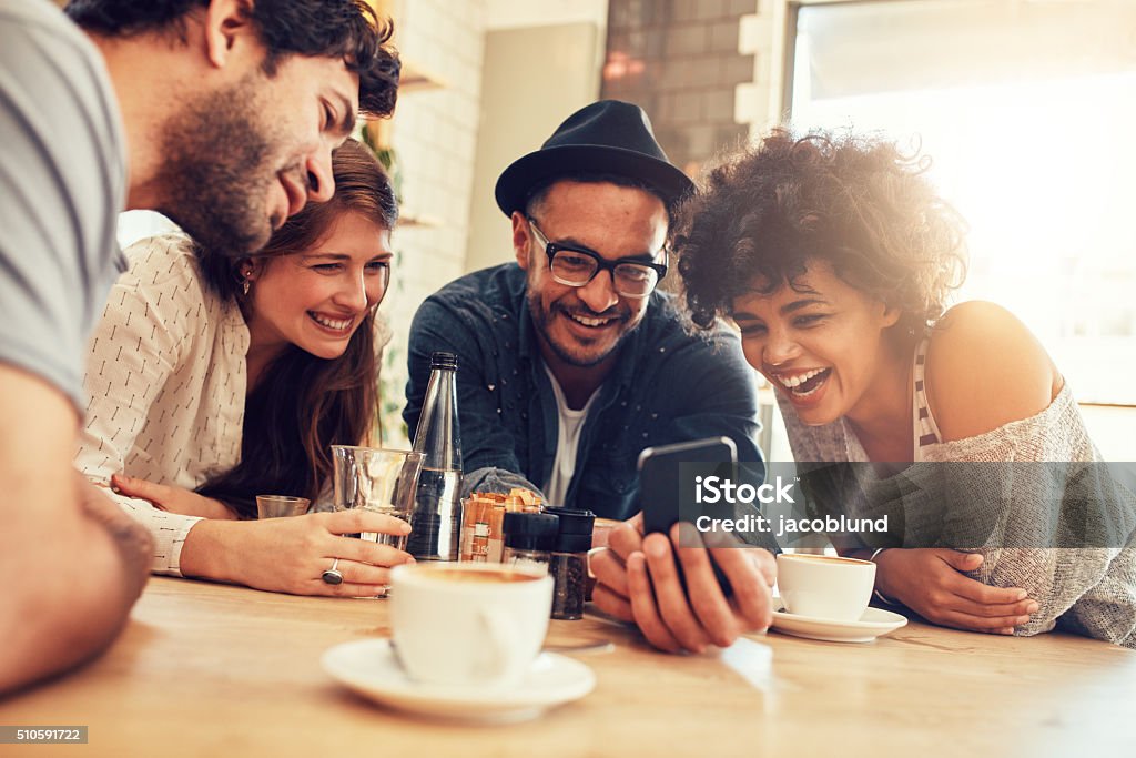 Friends looking at smart phone while sitting in cafe Portrait of cheerful young friends looking at smart phone while sitting in cafe. Mixed race people sitting at a table in restaurant using mobile phone. Friendship Stock Photo