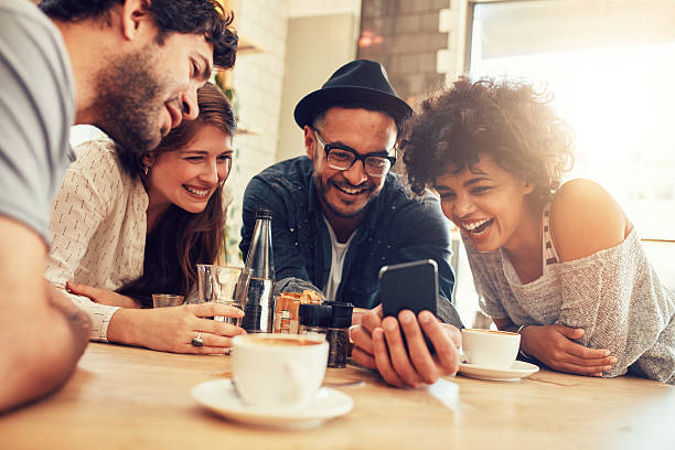 Friends looking at smart phone while sitting in cafe Portrait of cheerful young friends looking at smart phone while sitting in cafe. Mixed race people sitting at a table in restaurant using mobile phone. four people photos stock pictures, royalty-free photos & images