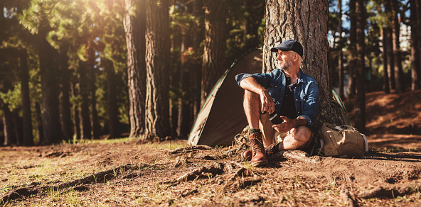 Portrait of senior man sitting by a tree with a tent in background. Mature man sitting at a campsite.