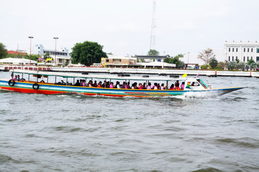 Bangkok, Thailand - February 19, 2013: Tourboat with tourists is on way upstream river Chao Praya in Bangkok. On boat is large goup of Asian people. Guide with microphone is standing in front of people. Three men are sitting in back alone.