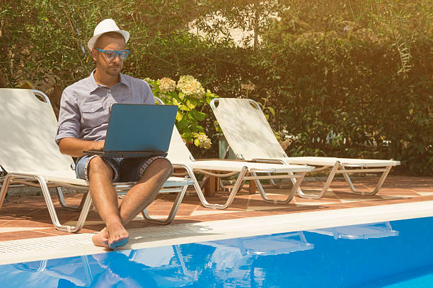 Young man sitting at the poolside and working on his laptop 