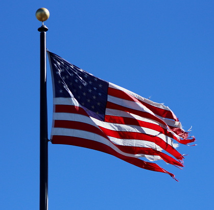 Frayed American flag waving against bright blue sky.