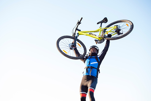 Smiling sportsman raising up his mountain bicycle on bright blue sky background.