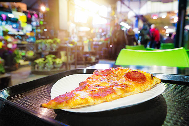 Pepperoni pizza slice on paper plate at market with sunshine in the background.