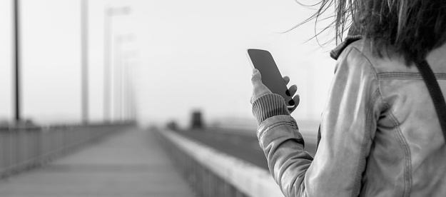 Picture of young woman standing on the bridge and using smartphone. She is wearing leather jacket.
