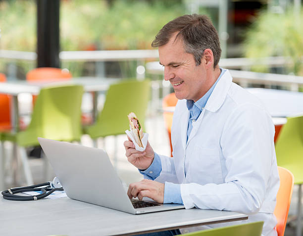Doctor working online at the hospital's cafeteria Happy doctor working online on a laptop computer at the hospital's cafeteria while having a bite cafeteria sandwich food healthy eating stock pictures, royalty-free photos & images