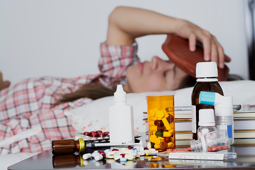 Woman lying in a bed, with a hot water bottle in her head feeling pain, and a nightstand full of drugs, capsules, pills, thermometer, syringe and several medications.