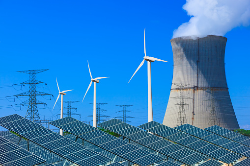 Solar panels with wind turbines in the foreground leading back to a atomic energy plant against a clear blue sky