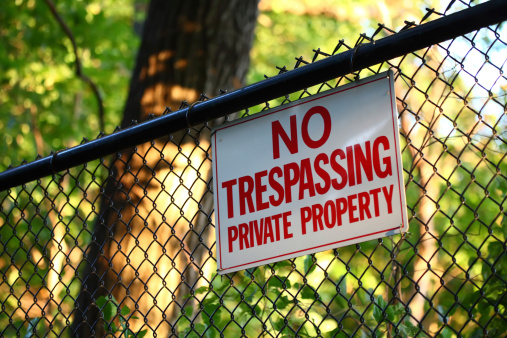 A close-up image of a 'No trespassing - private property' sign hanging on a fence with green trees on the background