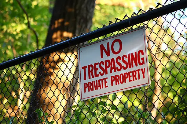 A close-up image of a 'No trespassing - private property' sign hanging on a fence with green trees on the background
