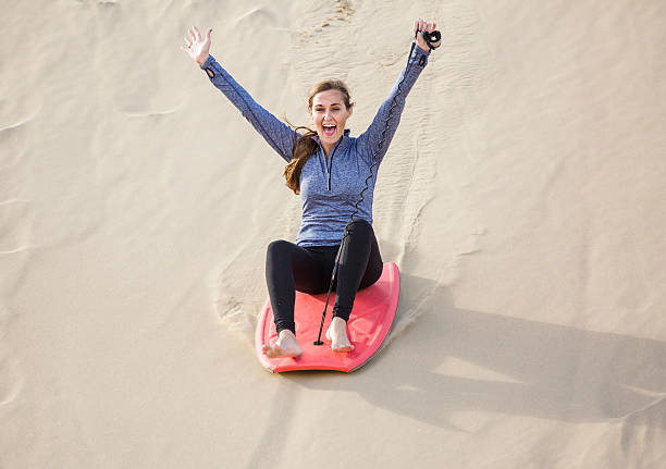 Young Woman Playing in the Sand Dunes Outdoor Lifestyle An excited and happy young woman riding a board down a sand dune hill having fun playing outdoors while on vacation sliding down stock pictures, royalty-free photos & images