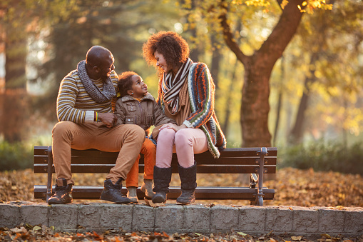 Smiling African American parents sitting with their daughter on a bench in nature and talking to each other.