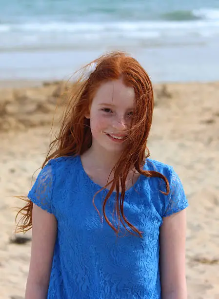 Photo showing a girl standing on a sandy beach, with the gentle sea waves lapping onto the shore in the background.  The girl is playing on the sand / pebbles as part of her summer seaside holiday, being pictured wearing a blue dress and a white flower in her long red hair.