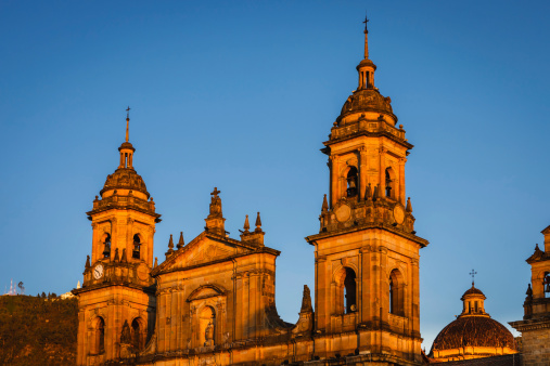 The rays of the setting sun hit the spires of the Catedral Primada, or translated into English, Primary Cathedral of Bogota, located on the East side of Plaza de Bolivar or Bolivar Square, in the Andean capital city of Bogota, in Colombia in South America. The sunlight turns the towers to gold.  Completed in 1823, the Cathedral is the seat of the Roman Catholic Archbishop of Colombia and a fine example of Colonial Spanish architecture. In the background is the Andean peak of Monserrate.  Photo shot in the evening sunlight against a clear blue sky; horizontal format; copy space.