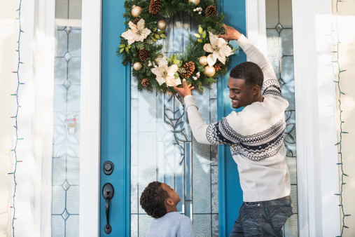 African American man (30s) hanging holiday wreath on front door.  He is looking down, talking to adorable son (5 years).  
