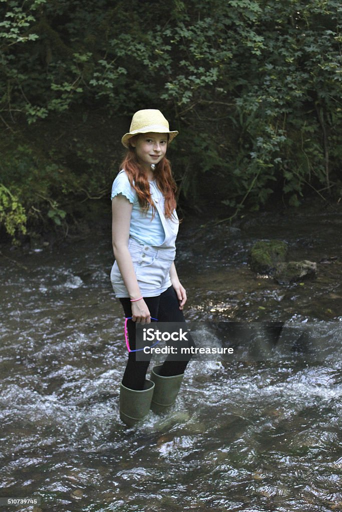 Image of girl playing, paddling, wading in river, woodland stream Photo showing a young girl with long hair playing / paddling / wading in a shallow river as part of a woodland walk to cool down on a particularly hot, sunny day. She is pictured wearing green wellies / wellie boots, demin dungaree jeans, pink sunglasses and a straw hat, and carefully walking in the water, making sure that it isn't too deep. Adolescence Stock Photo