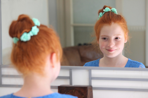 Photo showing a young girl with long ginger / red hair styled as a bun, pictured wearing a string of artificial turquoise flowers (blue daisy chain) and looking at her reflection in a mirror, posing to herself.