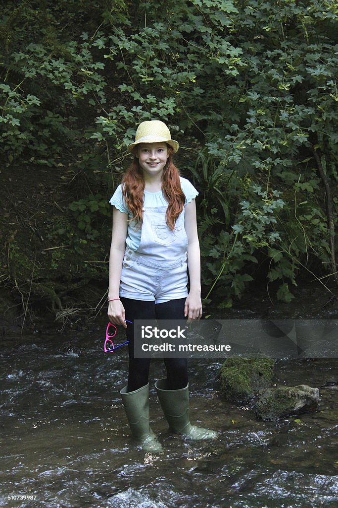 Image of girl playing, paddling, splashing, wading in river, woodland Photo showing a young girl with long hair playing / paddling / splashing / wading in a shallow river as part of a woodland walk to cool down on a particularly hot, sunny day. She is pictured wearing green wellies / wellie boots, denim dungaree jeans and a straw hat, holding her pink plastic sunglasses and carefully walking in the water, making sure that it isn't too deep.  As she wades through the river, she passes through pockets of shade and strong light / rays of sunshine, which lights her up as if she is standing in a natural spotlight. Girls Stock Photo
