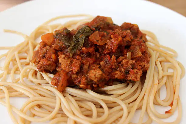 Photo showing a plate of homemade spaghetti bolognese, served on a white plate with the rich bolognese sauce piled high in the centre of the spaghetti pasta.  This traditional Italian dish is being served up as a family meal at teatime.