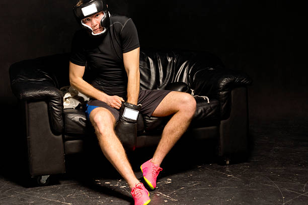 Young boxer fitting his boxing gloves as he prepares himself for a fight sitting on a black leather couch on a dark background