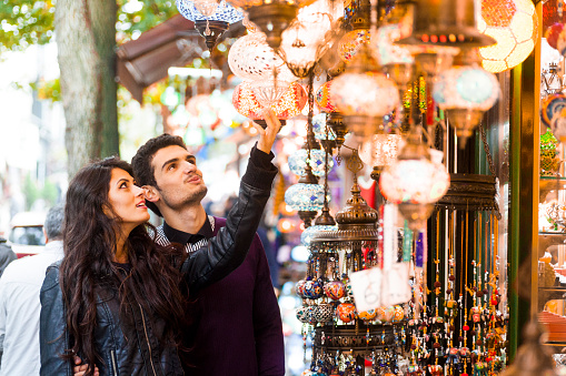 Young couple of adults is admiring the lamps sold in the street market. The girl is checking the lamps by touching them and rotating while the guy is making an impressed face. The image is colourful and warm, has a magical feel to it. Copy space available. Made in Istanbul, Turkey.