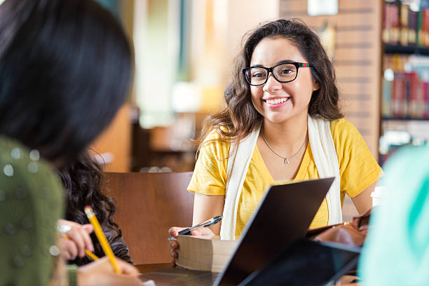 Trendy college students hanging out in library while studying Trendy hipster Hispanic college age girl is smiling while sitting at tabe in library with group of friends. Students are working in study group to prepare for exam while using laptop computers, digital tablets, and other technology. group of people wearing eyeglasses stock pictures, royalty-free photos & images