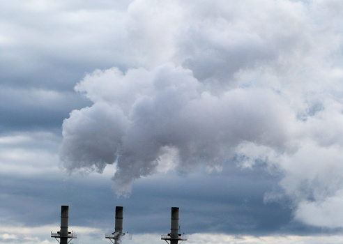 Three industrial steam chimneys with billowing smoke on a blue cloudy sky.  The industry stacks are a concern of environmental pollution.