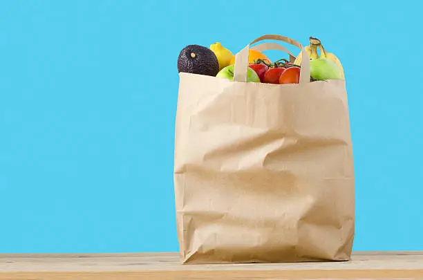 A brown paper shopping bag, filled to the top with varieties of fruit, on a light wood surface.  Isolated on a turquoise blue background.