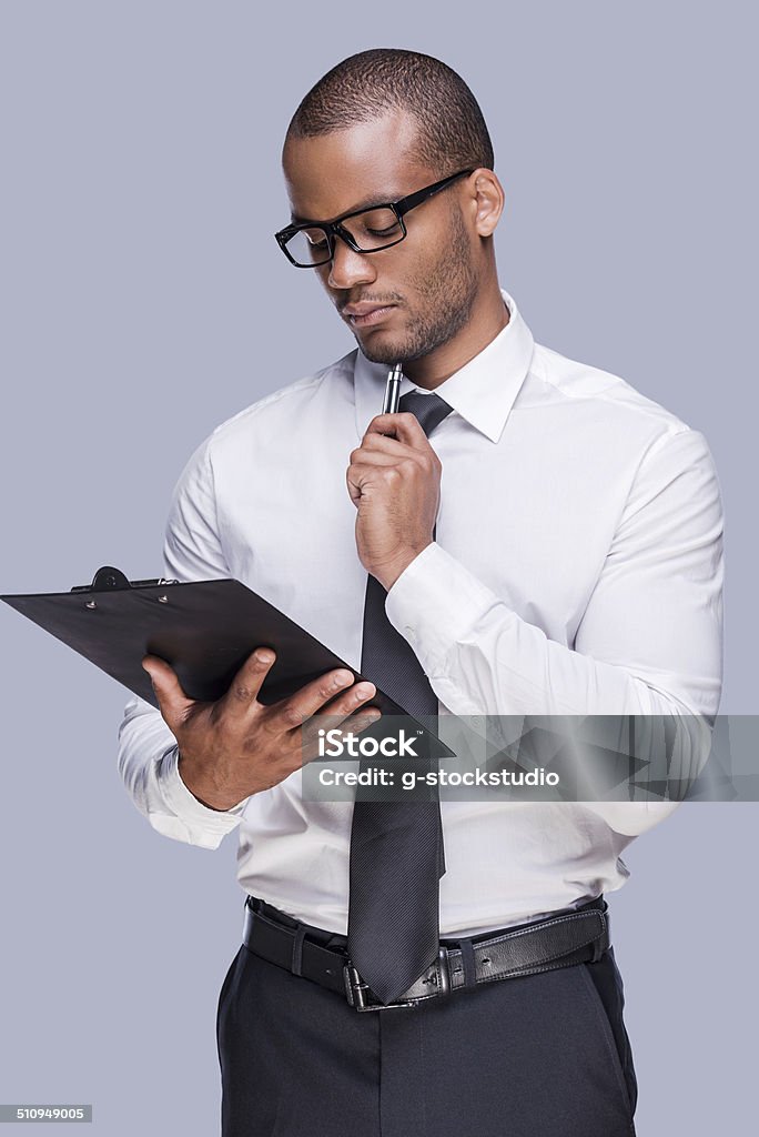 Examining contract before signing. Thoughtful young African man in shirt and tie holding clipboard and touching his chin with pen while standing against grey background Clipboard Stock Photo