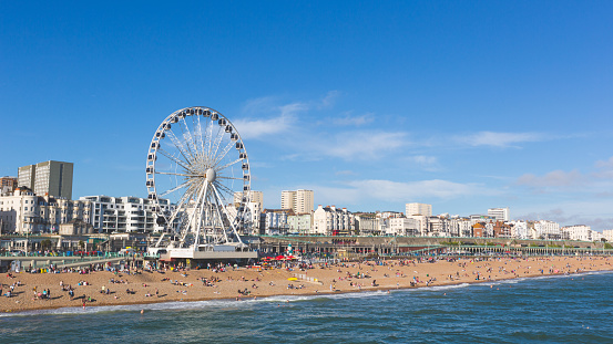 Brighton view of seaside from the pier. Panoramic shot with the famous ferris wheel, the stones beach with unrecognizable persons on a sunny summer day.