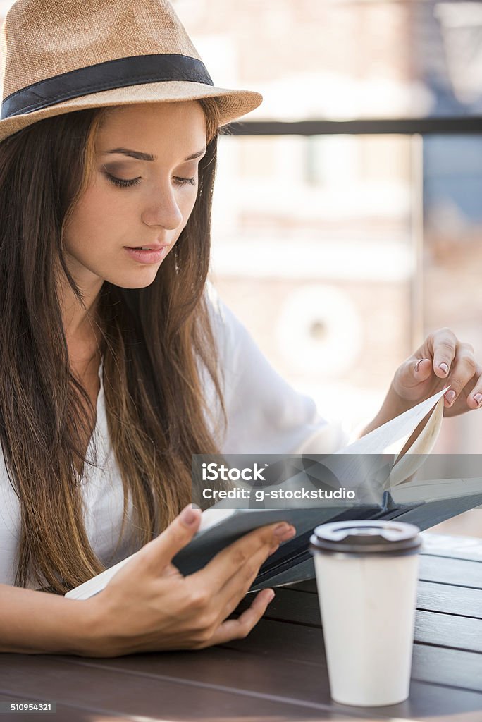 Reading her favorite book. Beautiful young woman in funky hat reading book and smiling while sitting outdoors Adult Stock Photo