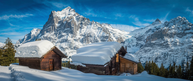 Grindelwald, Switzerland - 25th February 2014: Traditional wooden chalets high in the Alps buried under crisp white snow below mountain peaks. Composite panoramic image created from five contemporaneous sequential photographs.