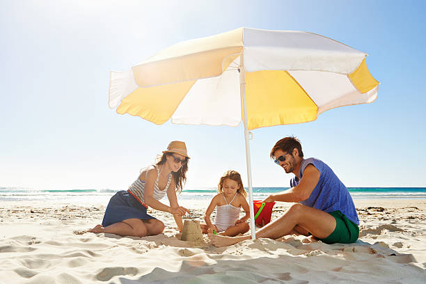 Helping their little girl build a sandcastle Shot of a young family building a sandcastle under an umbrella beach umbrella stock pictures, royalty-free photos & images