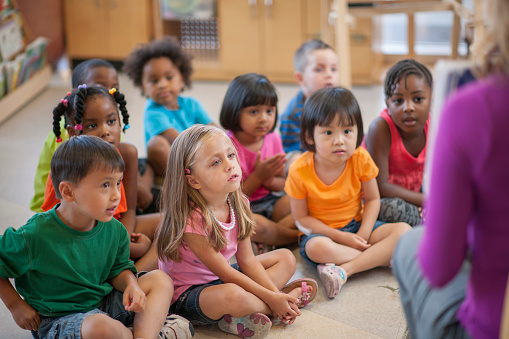 A multi-ethnic group of elementary age children are sitting on the floor and are listening to their teacher read them a story.