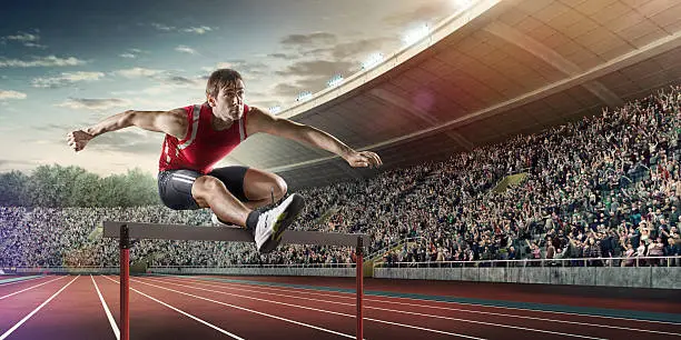 :biggrin:A professional male athlete jumping over a hurdle during a race. The action takes place in a outdoor . stadium full of spectators. The athlete is wearing generic athletics kit. Low angle image.