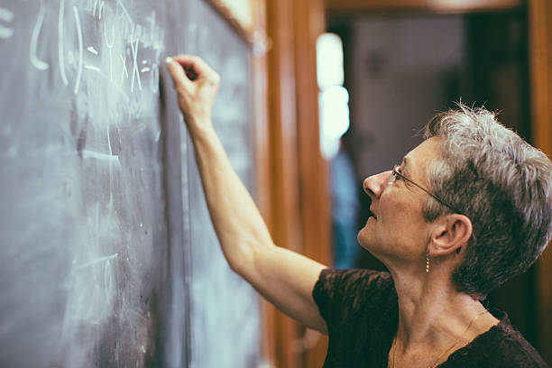 Mathemathics professor at chalkboard writing formula Concentrated senior female mathemathics professor writing a formula to the chalkboard.. Personal Perspective, Selective focus, small DOF. Natural interior light. leanincollection stock pictures, royalty-free photos & images
