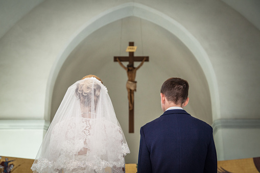 Photo taken during the wedding ceremony in Catholic church. Groom and bride stand before crucifix. Focus is on the crucifix. Newlyweds are in focus. Bride dressed in wedding dress and bridal veil. Groom dressed in suit and shirt. Photo taken from the back of bride and groom.