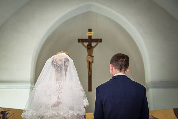 Bride and groom stand before crucifix in church 2 Photo taken during the wedding ceremony in Catholic church. Groom and bride stand before crucifix. Focus is on the crucifix. Newlyweds are in focus. Bride dressed in wedding dress and bridal veil. Groom dressed in suit and shirt. Photo taken from the back of bride and groom. catholicism stock pictures, royalty-free photos & images