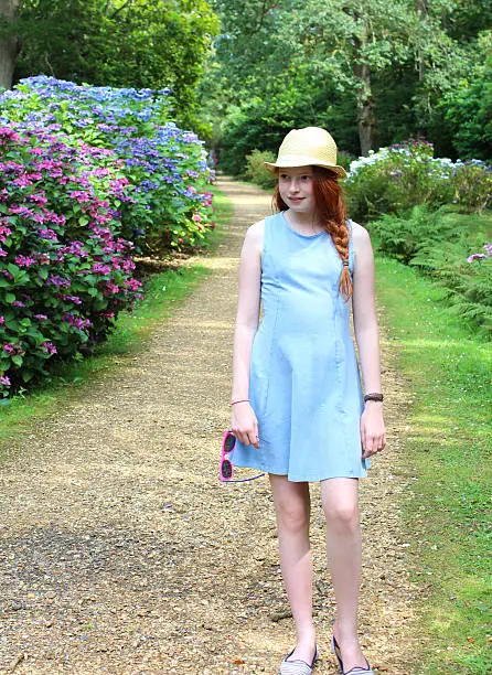 Photo showing a young girl, with long red hair, a blue dress and a straw hat, walking past a mass of lilac and pale blue flowers growing on a large hydrangea bushes (Hydrangea macrophylla) next to the garden path.  The mophead and lacecap hydrangeas are enjoying a shady part of the woodland garden, beneath some large trees that cast shade, and are pictured flowering in the middle of the summer.