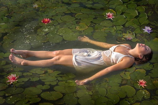 In the classic style based on Shakespeare's Ophelia, a beautiful young lady lays motionless in a pool of water wearing a white dress and surrounded by waterlilies. Inspired by the Pre-Raphaelite John Everett Millais. A great image to depict this movement or the Art Nouveau period. 