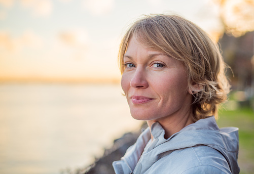 Blonde, short hair, blue eyed, mid 30's woman gives a strong contented smile as the sun sets behind her.