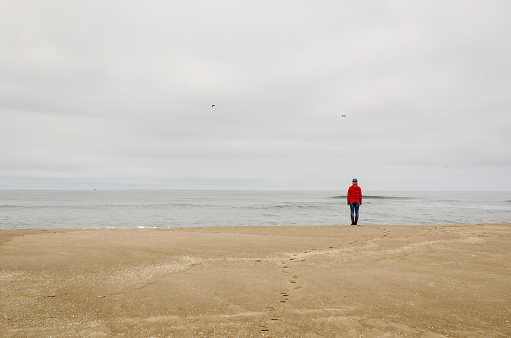 A woman in a red jacket and blue cap stands on the beach alone.  She is looking off in the distance in this calm scene.