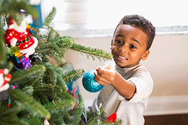 Adorable African American boy (5 years) hanging ornament on Christmas tree.  He is smiling, looking for a branch to hang the blue ornament that he is holding in his hand, reaching up.