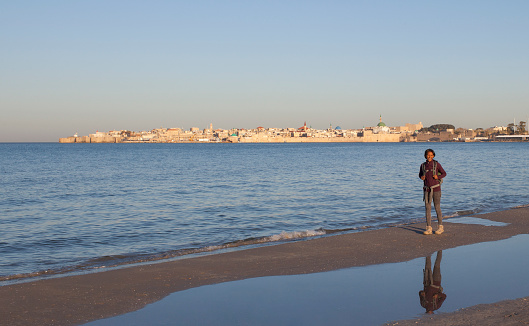 Women’s adventure travel. Female backpacker traveling in Akko, Israel. Tourist woman standing on beach of Mediterranean sea bay in front of landscape with view on Acre old city.