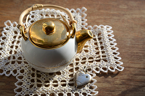 Tea time:porcelain teapot on a lacy napkin with decorative silver heart on wooden background.