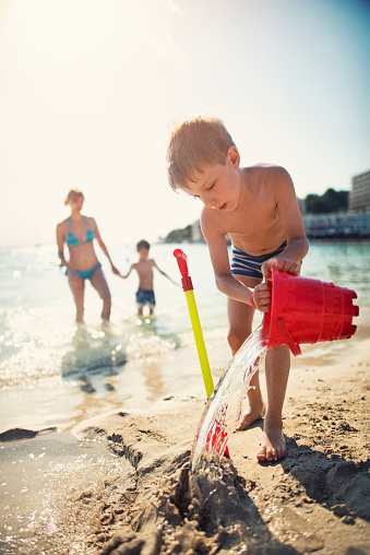 Little boy having fun playing on the beach. The boy is pouring water and preparing the spot for sandcastle. The summer sun is flooding the beach with warm light. Mother and brother is visible in the background.