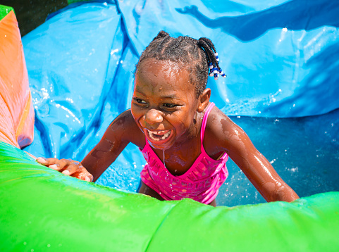 Cute smiling little African American girl playing on an inflatable bounce house outdoors. Having fun playing on a bounce house water slide at an outdoor party