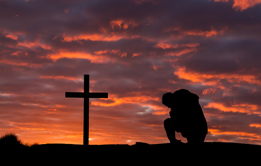 Man praying by a cross as the setting sun light up the clouds.