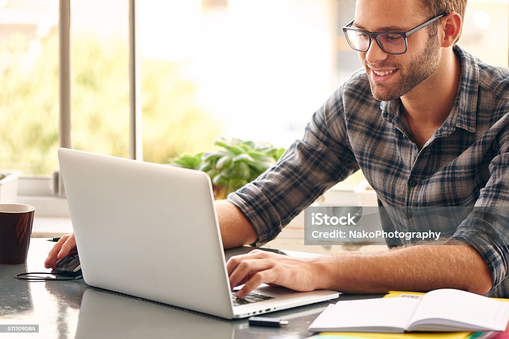 Happy man sitting and working at his desk from home Happy young man, wearing glasses and smiling, as he works on his laptop to get all his business done early in the morning with his cup of coffee Adult Stock Photo