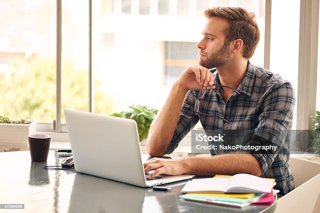 Young entrepreneur looking out the window in thought Young man sitting in front of his notebook with a cup of coffee, looking off to the side with his glasses in his hand, as he considers what to write next in his paragraph Adult Stock Photo